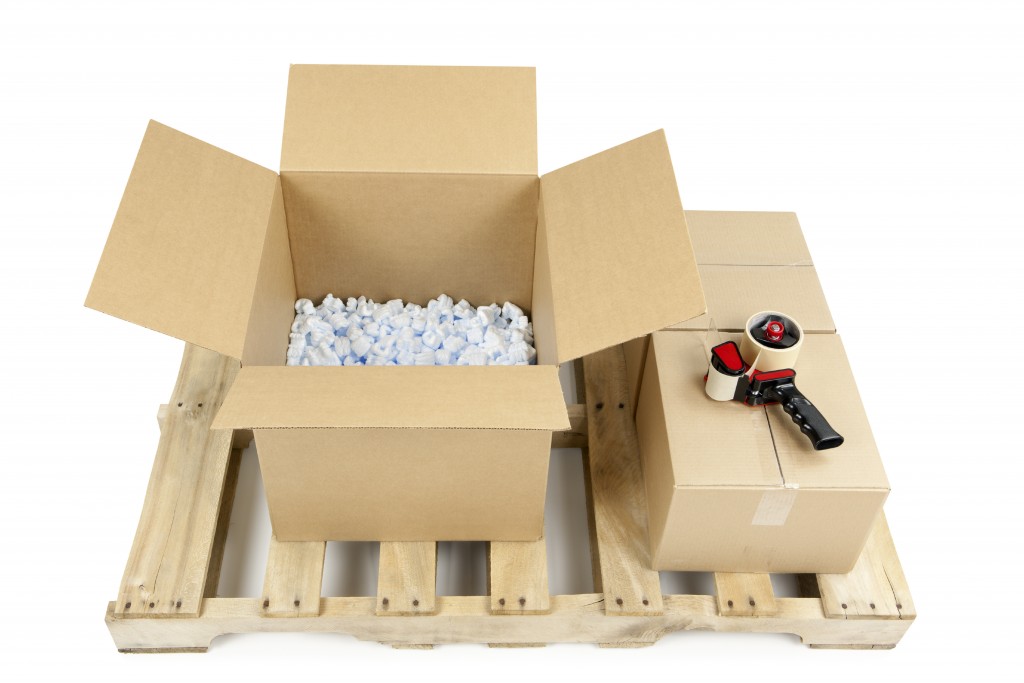 Shipping pallet with one new open cardboard box half full of packing peanuts, and two closed boxes with a tape gun. Isolated on white.